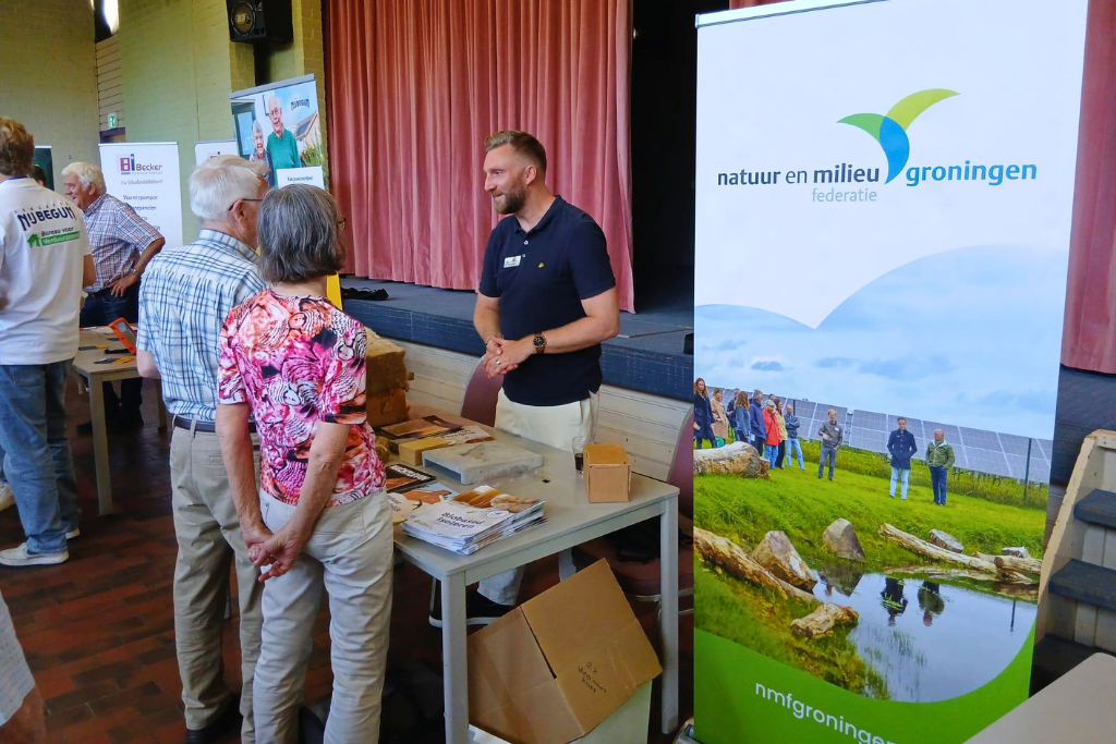 Merijn de Jong van de Natuur en Milieufederatie die naast een banner en achter een tafel met een natuurinclusief zonnepark staat te praten met een ouder echtpaar.