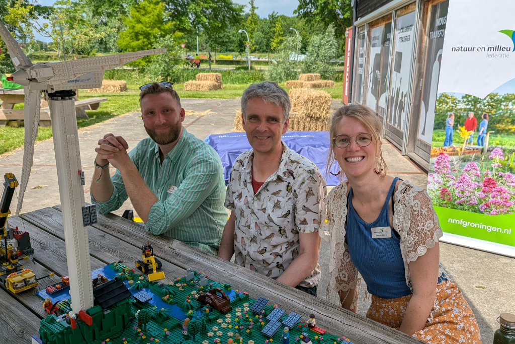 Drie collega's van de NMG die lachend op een picknick tafel zitten met voor hun een Lego-energietuin met daarop zonnepanelen, een windmolen en heel veel ecologie.