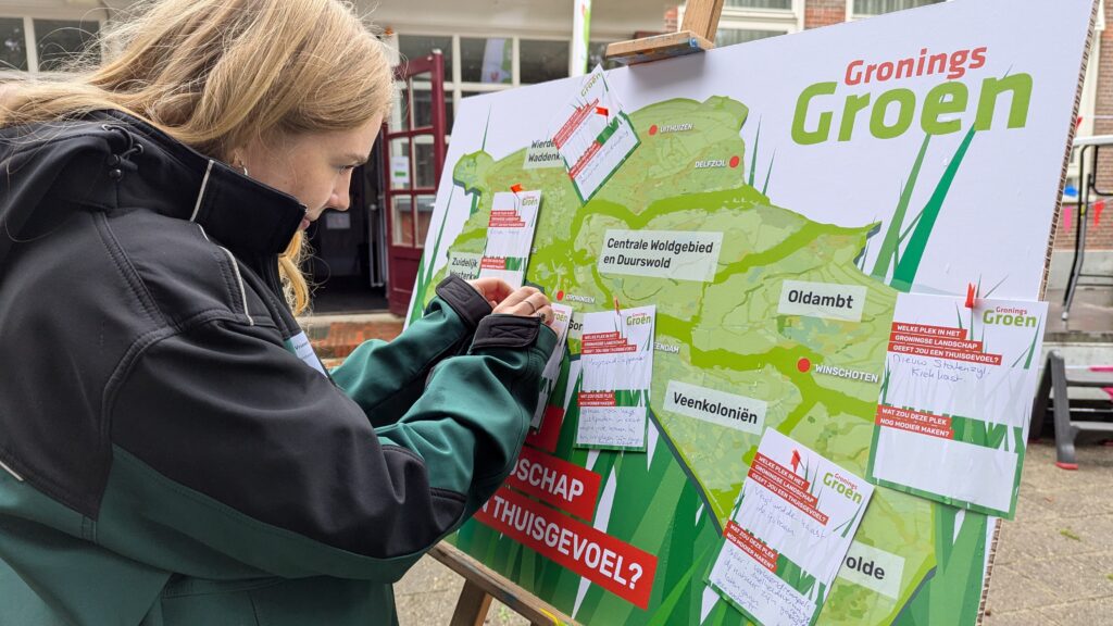 Jonge blonde vrouw die een papiertje op een bord met de vraag: ‘Welk landschap geeft jou een thuisgevoel?’ prikt met een punaise.